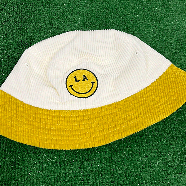 LA Be Happy smiley face embroidered on white corduroy yellow brim bucket hat on grass background - Free & Easy